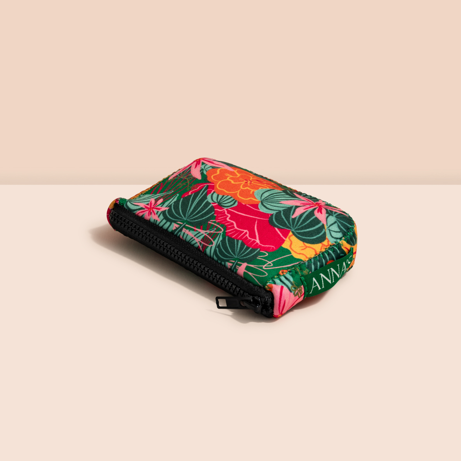 Angled side-profile view of a compact tropical pattern cosmetic travel pouch showing the black zipper closure and green Anna's brand tab.