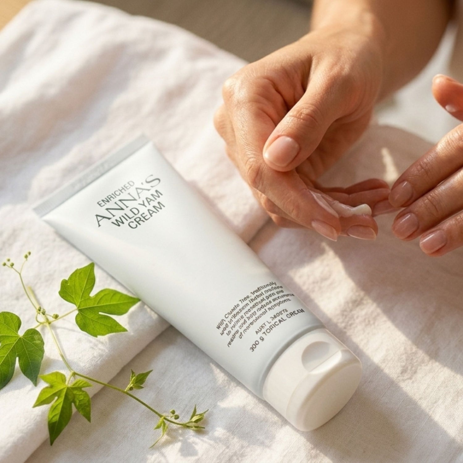 A close-up of a person rubbing a white cream onto their fingertips, next to a tube of Enriched Anna's Wild Yam Cream resting on a textured white cloth.