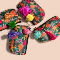 Overhead flat-lay of four colorful green tropical floral cosmetic bags with vibrant pink, purple, green, and yellow tassels.