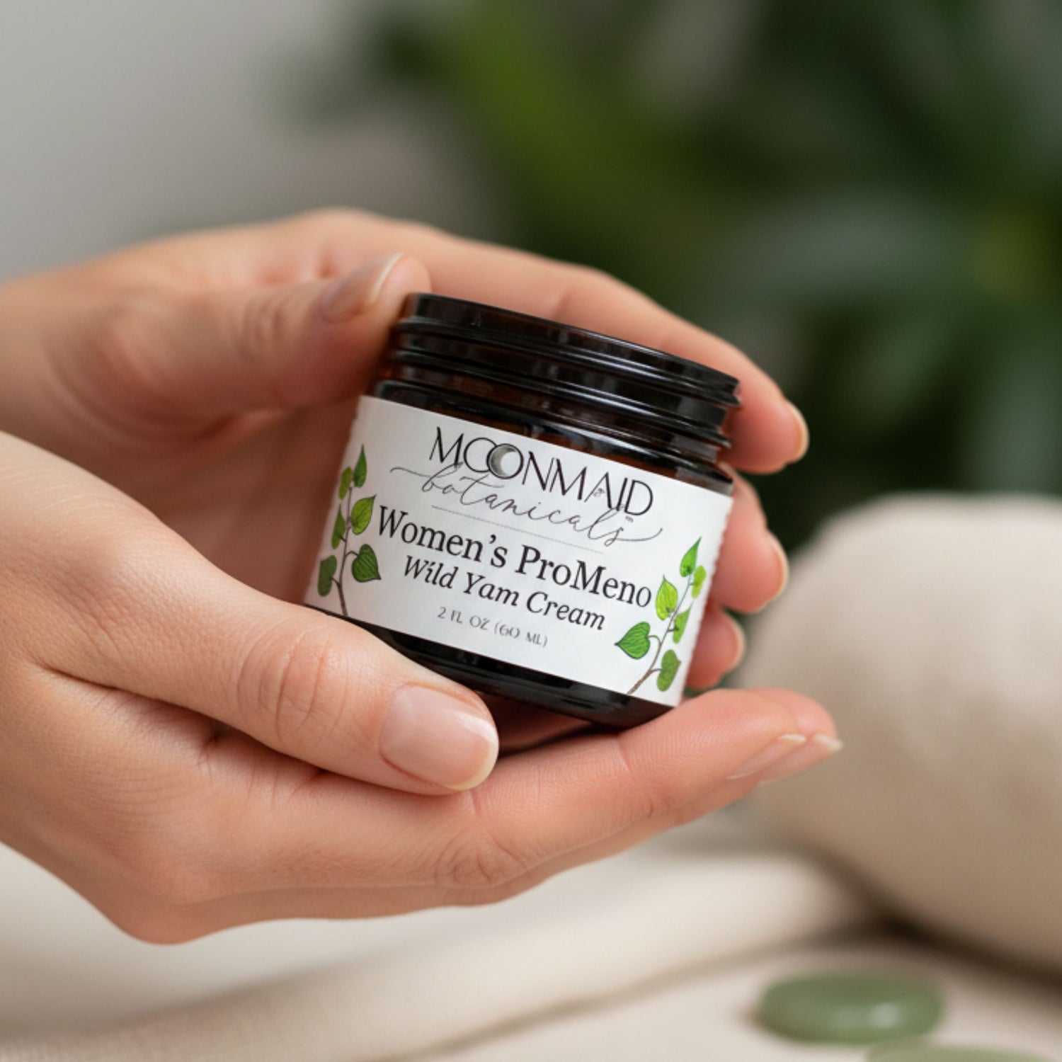 A close-up of a person holding a small amber glass jar of MoonMaid Botanicals Women's ProMeno Wild Yam Cream in both hands.