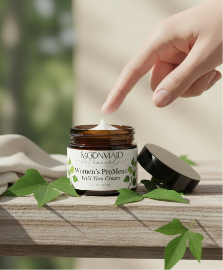 An open amber glass jar of Moonmaid Botanicals Women's ProMeno Wild Yam Cream sitting on a wooden ledge surrounded by green botanical leaves.