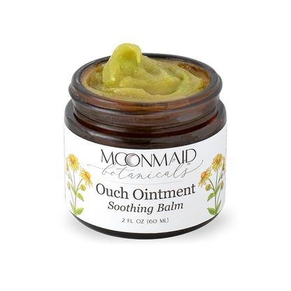 An angled close-up view of an open jar of Moonmaid Botanicals Ouch Ointment, highlighting its thick, natural green herbal salve texture.