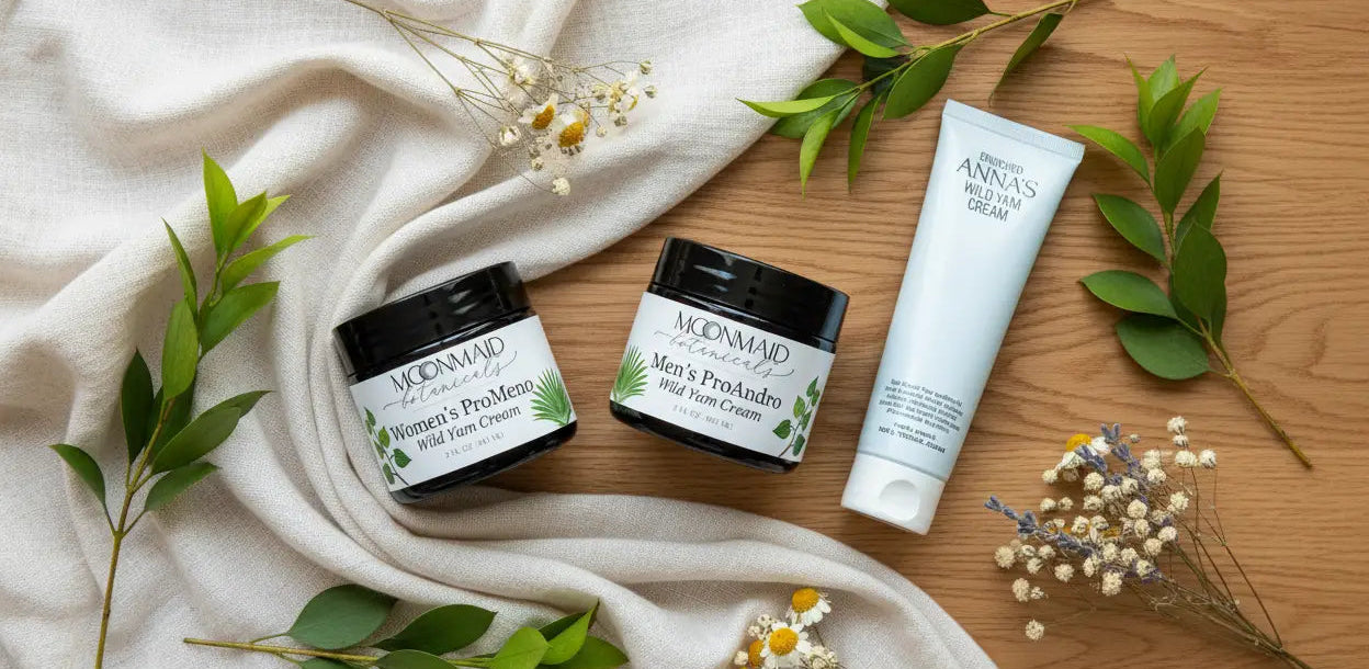 An overhead view of Moonmaid Botanicals Women's ProMeno and Men's ProAndro wild yam creams next to Anna's Wild Yam Cream tube, arranged on a neutral fabric background with green leaves and dried flowers.